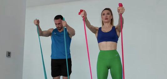 Man and woman doing resistance band front raise for front delts at home – Bir erkek ve bir kadın evde ön omuz kasları için resistance band front raise hareketi yapıyor.